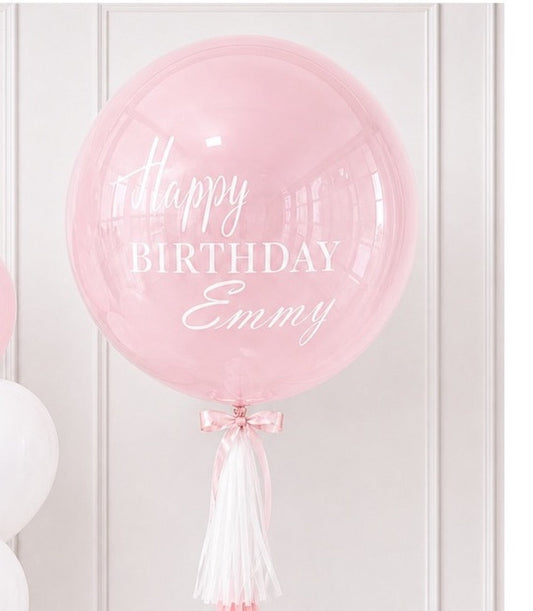 Pink clear bubble balloon with "Happy Birthday Emmy" in white text, decorated with pink bow and white tassel tail.
