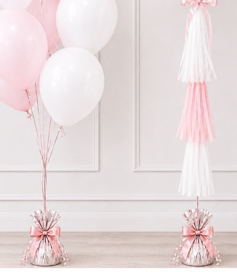 Pink and white balloon bouquets with tassel garlands and ribbon bows on wooden floor against white paneled wall.