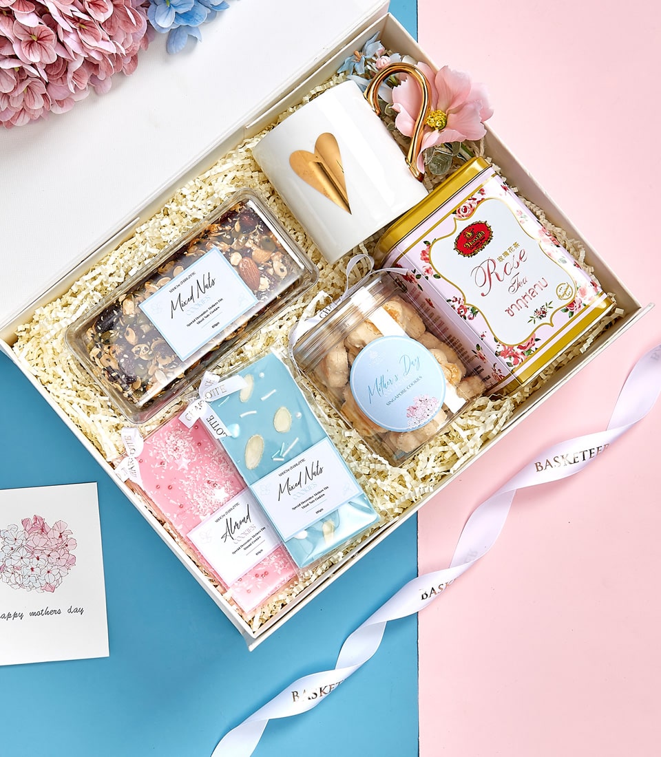 Elegant Mother's Day gift box with gold heart mug, rose tea, granola, cookies, and chocolate bars on blue and pink background
