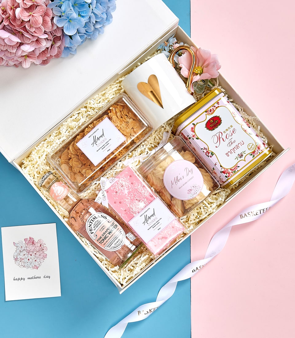 Mother's Day gift set with gold-heart mug, tea tin, cookies, and pink grapefruit tonic on blue-pink background