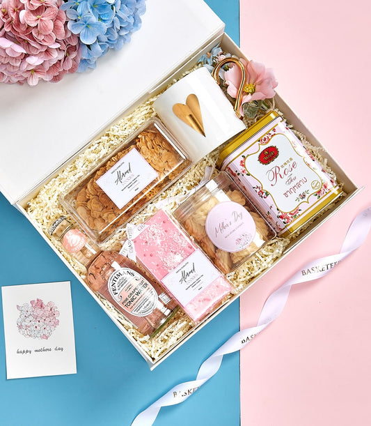 Mother's Day gift set with gold-heart mug, tea tin, cookies, and pink grapefruit tonic on blue-pink background