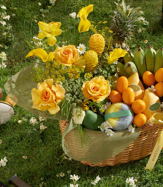 Woven wicker gift basket overflowing with golden roses, tropical fruits, and yellow ginger flowers on lush green garden