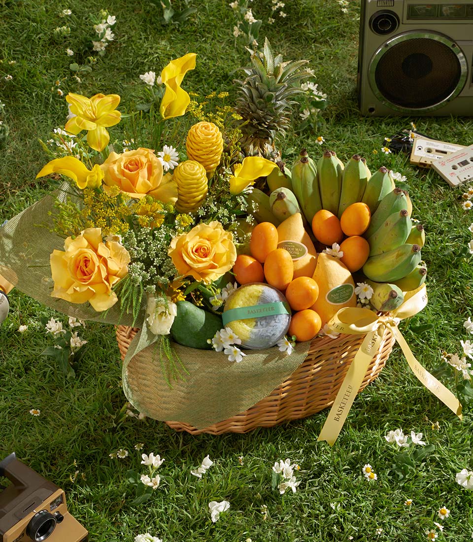Large wicker gift basket with fresh tropical fruits, yellow roses, and calla lilies on green lawn