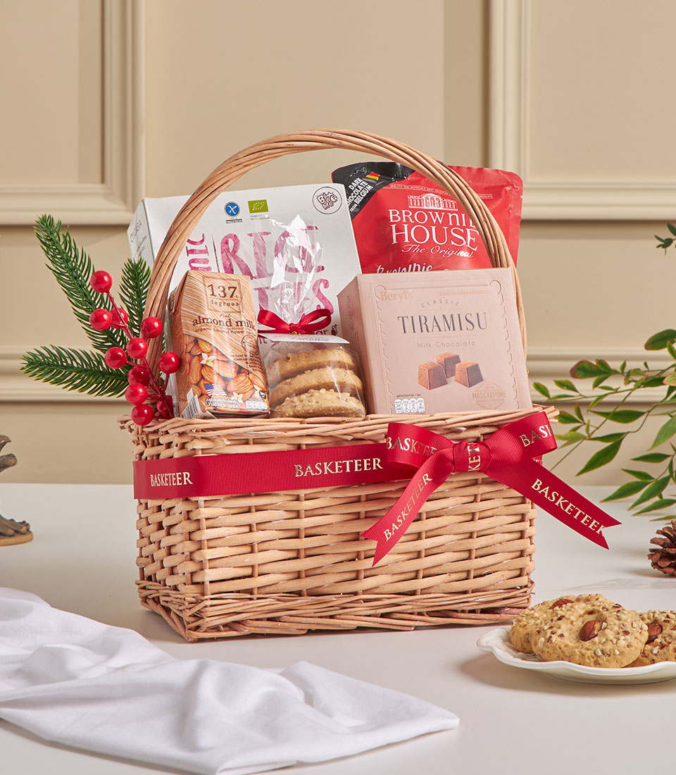 Woven wicker gift basket with tiramisu chocolate, almond milk, cookies and brownie mix, red ribbon bow, festive pine