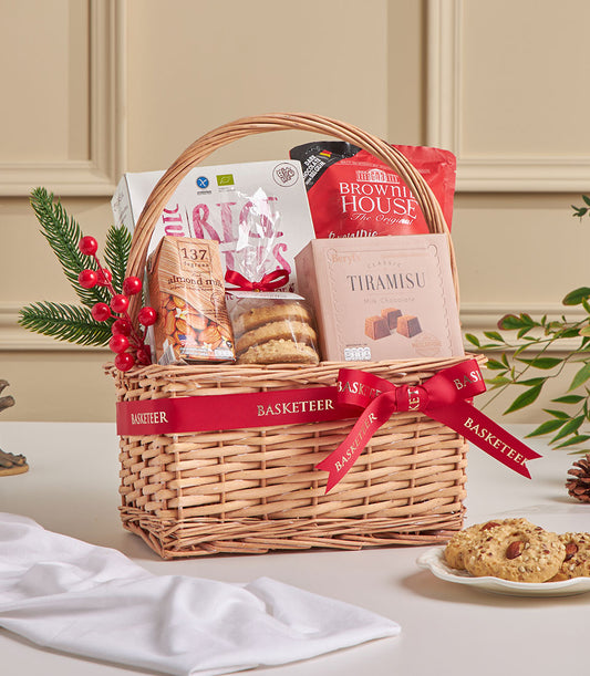 Woven wicker gift basket with tiramisu chocolate, almond milk, cookies and brownie mix, red ribbon bow, festive pine