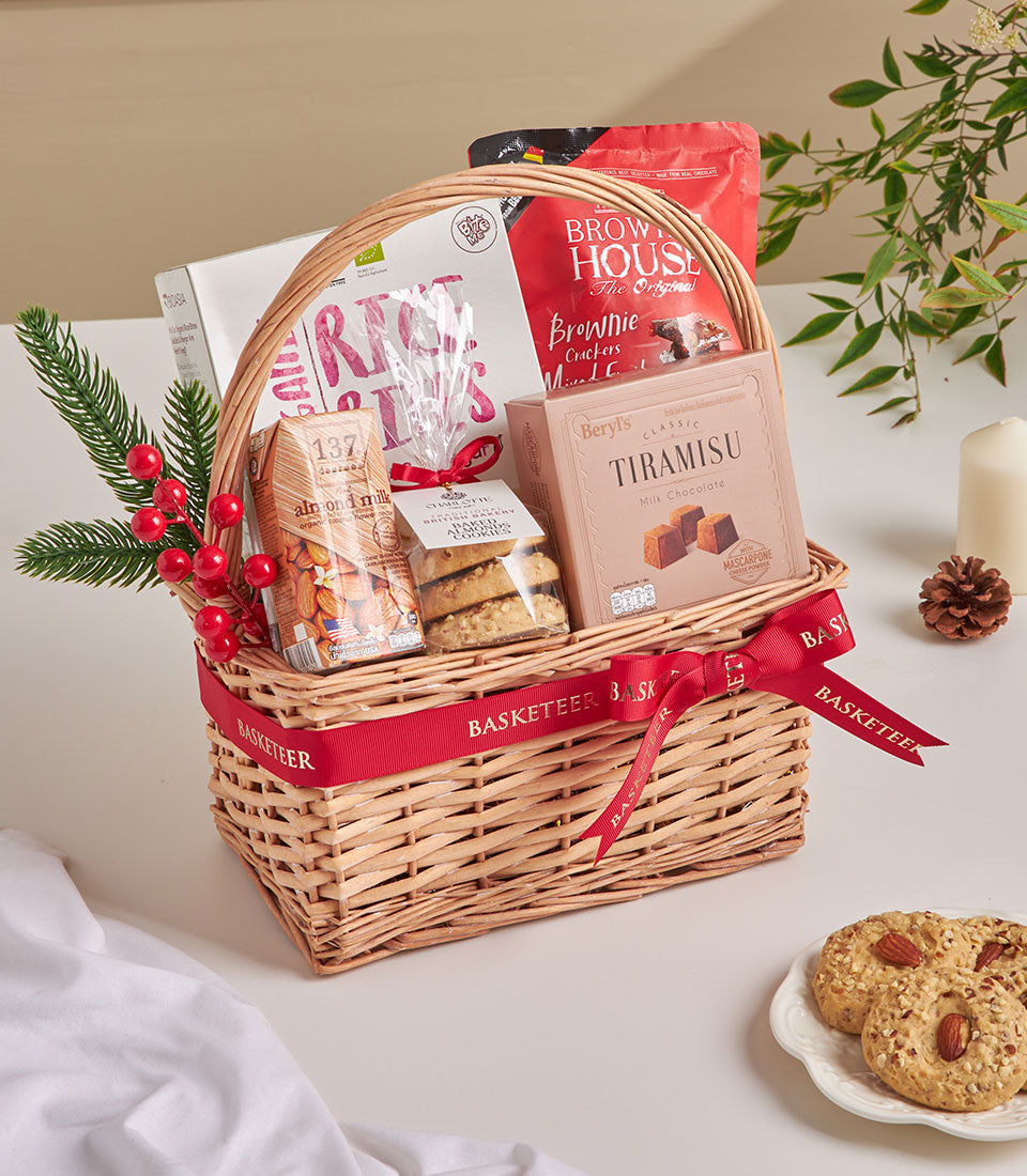 Wicker gift basket with tiramisu chocolate, almond milk, brownie crackers, and mini cookies tied with red ribbon, styled for