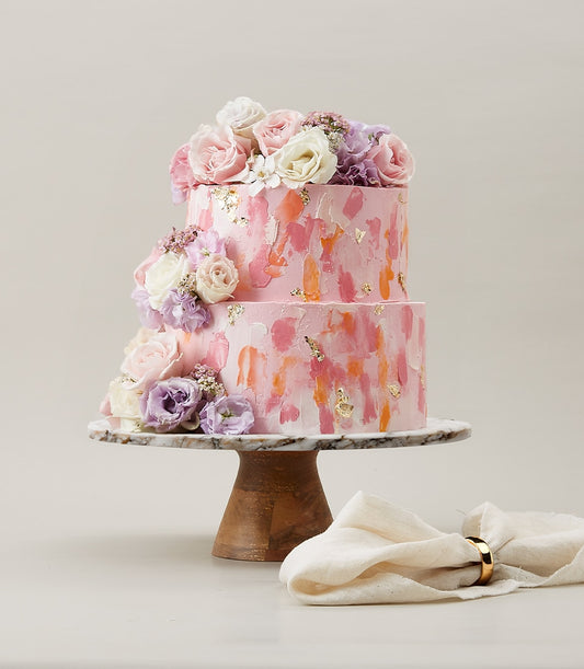 Blush two-tier birthday cake with painterly pink frosting, fresh flowers, and gold leaf accents