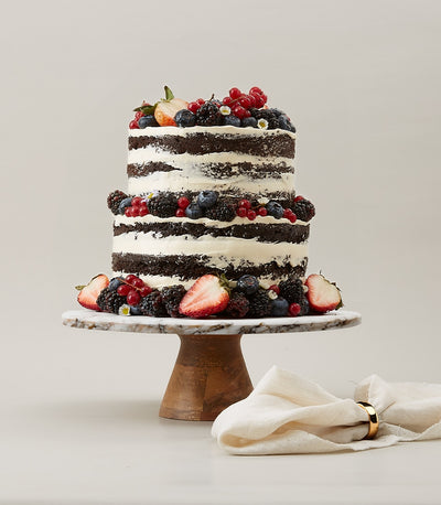 Two-tier naked chocolate cake with white buttercream, fresh strawberries, blueberries, and blackberries on marble stand