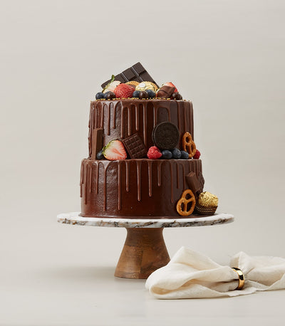 Two-tier chocolate drip cake with berries, chocolate bars, Oreos, and gold Ferrero Rocher on marble stand