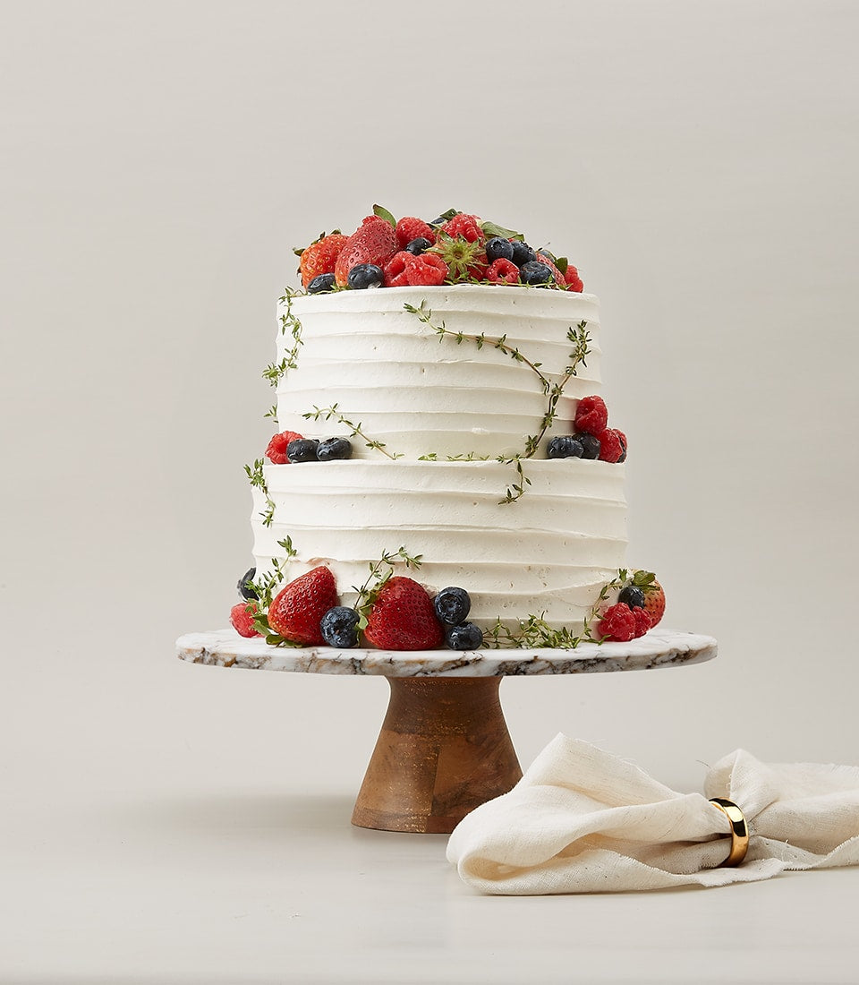 Two-tier white vanilla cake with fresh strawberries, raspberries, and blueberries on marble stand