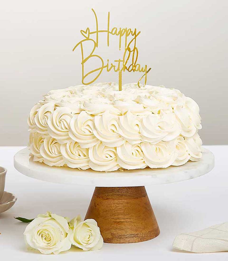 White layer cake with rosette frosting and gold Happy Birthday topper on marble stand with roses