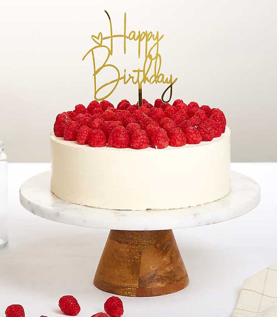 White birthday cake with fresh raspberries and gold Happy Birthday topper on marble stand