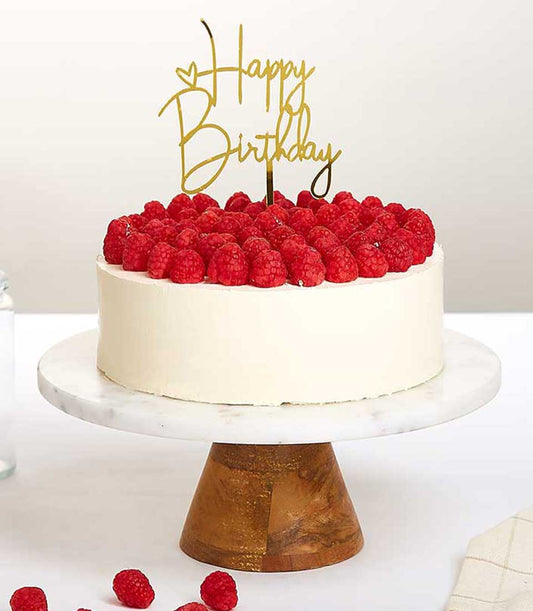 White birthday cake with fresh raspberries and gold Happy Birthday topper on marble stand