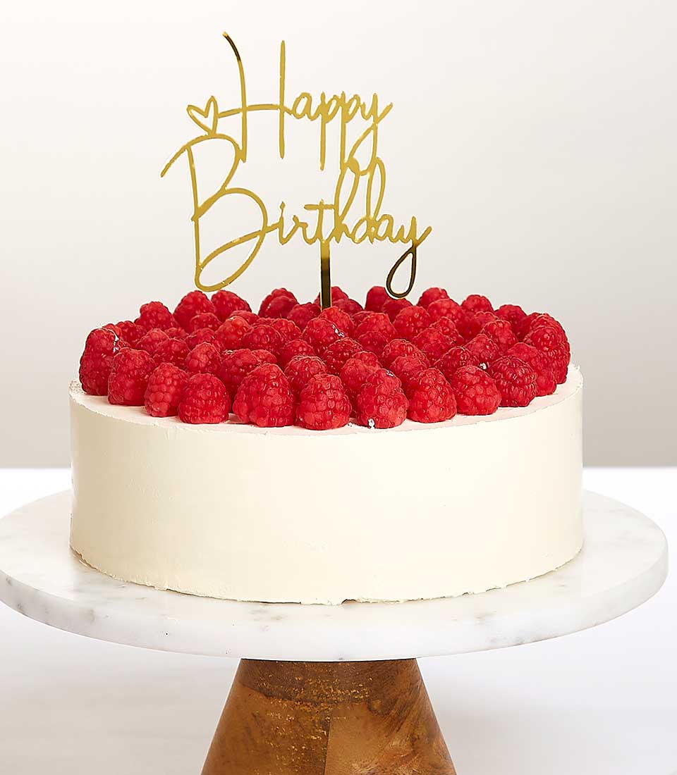 White frosted birthday cake with fresh raspberries and gold Happy Birthday topper on marble stand