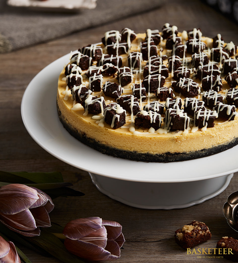 Brownie cheesecake with dark crust, creamy filling, and fudgy brownie topping on ceramic stand with purple tulips