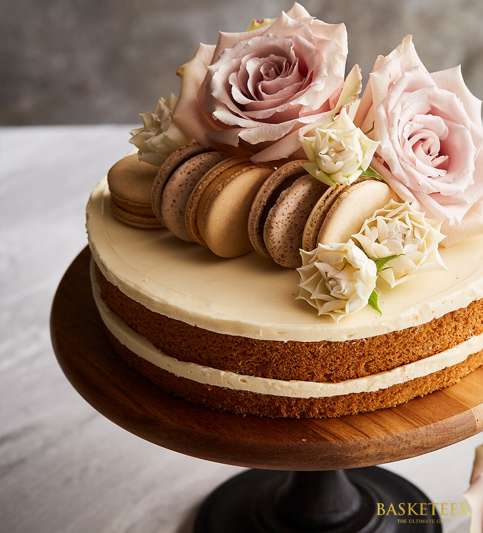 Two-tier vanilla butter cake with white frosting, pink and white roses, and macarons on wooden stand