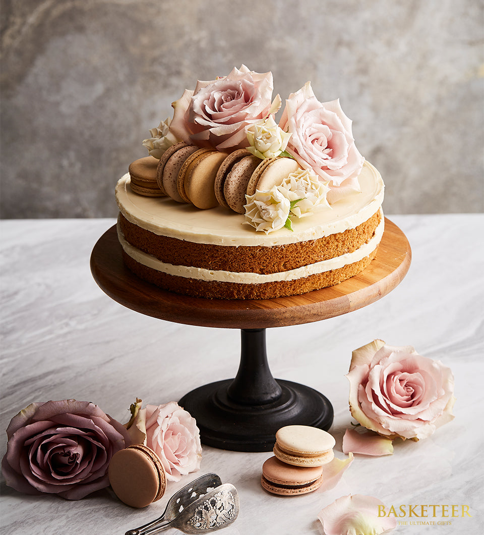 Vanilla butter cake with white frosting, pink and white roses, and macarons on dark wooden stand