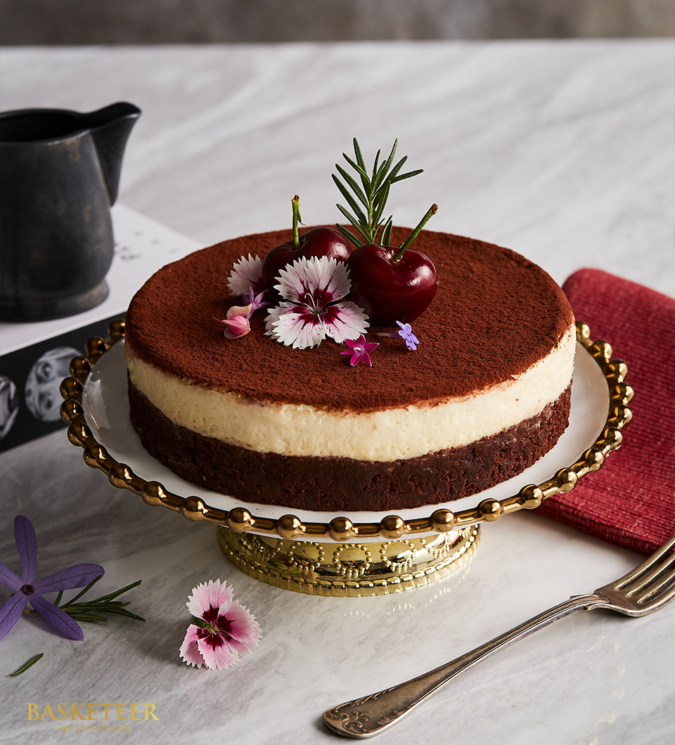 Rich chocolate layer cake with white cream and cocoa dust, garnished with cherries and edible flowers on gold stand