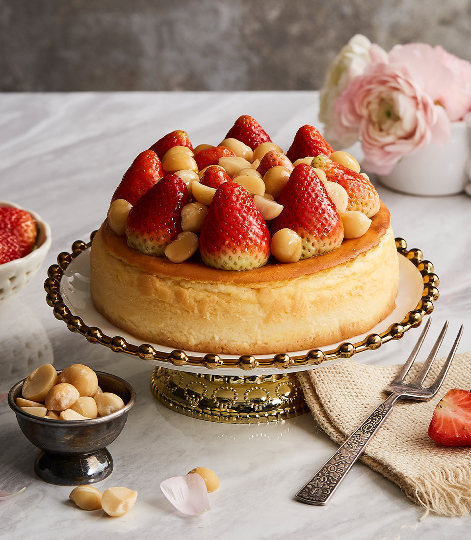 Strawberry macadamia cheesecake with golden crust on white cake stand, fresh berries and nuts, artisan birthday gift