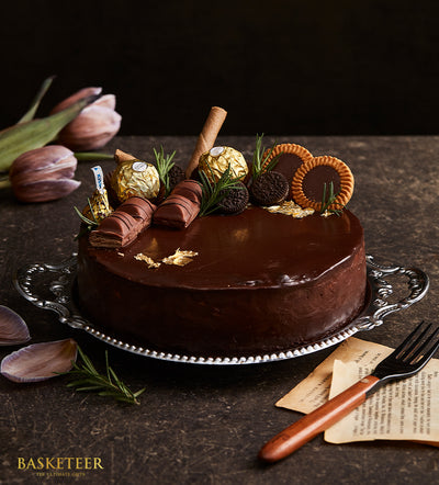 Decadent chocolate ganache cake with gold leaf, Kinder bars, and Kisses chocolates on silver platter, surrounded by fresh
