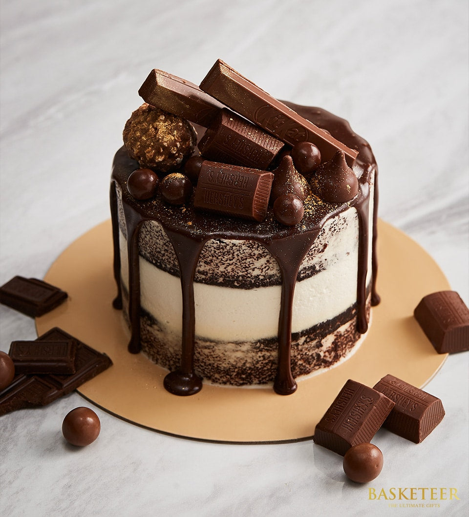 Semi-naked mini chocolate cake with white frosting, ganache drip, and assorted chocolate bar toppings on gold board