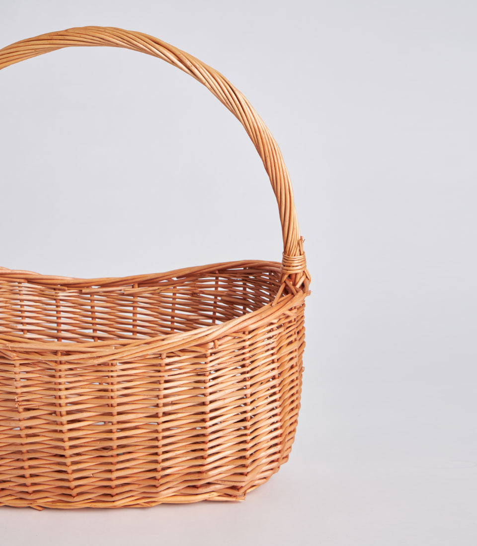 Light brown handwoven oval wicker basket with tall arched handle, natural rustic willow weave