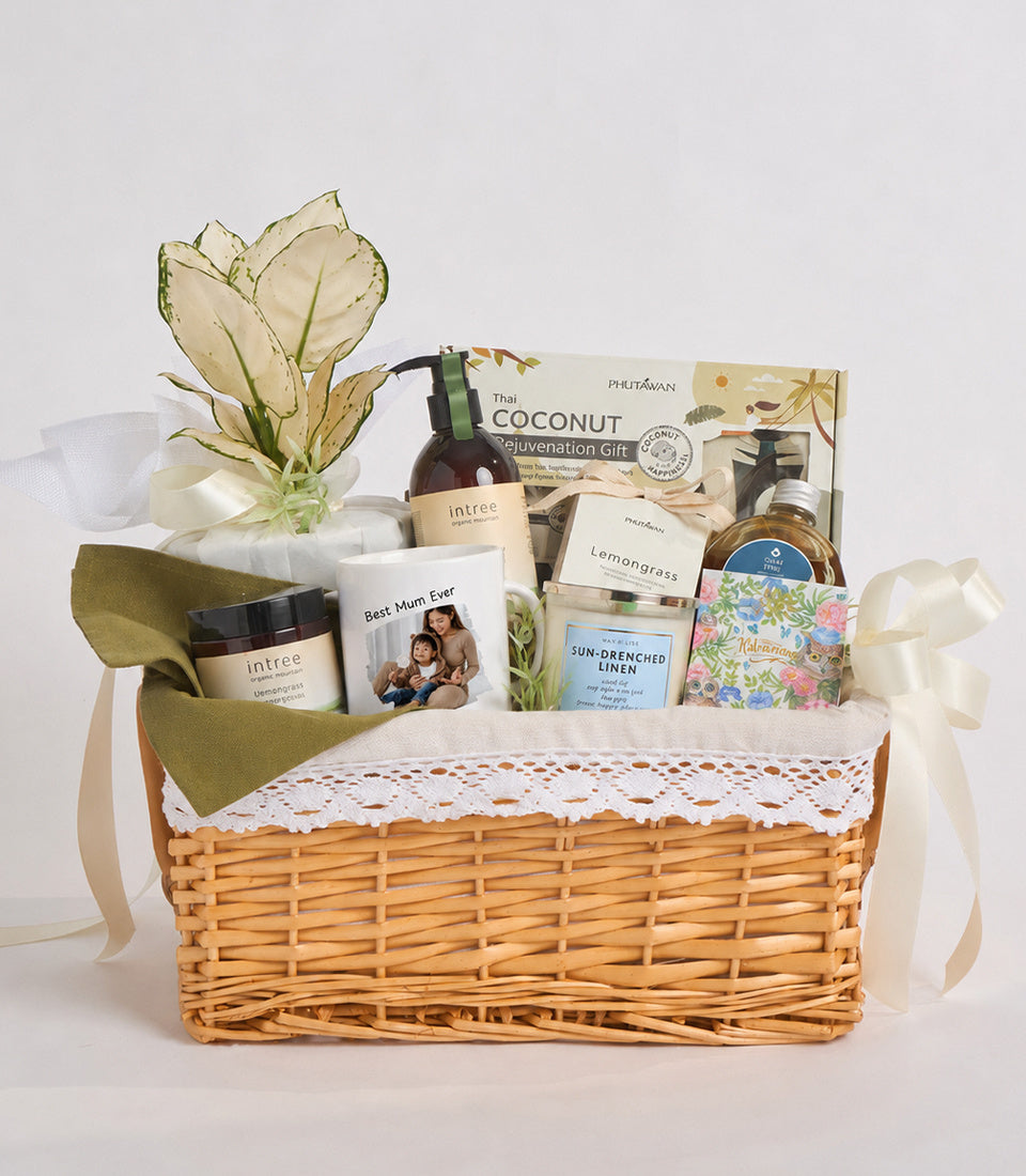 Wicker basket gift set with cream linens, skincare products, oils, candles, soap, and dried flowers on white background.