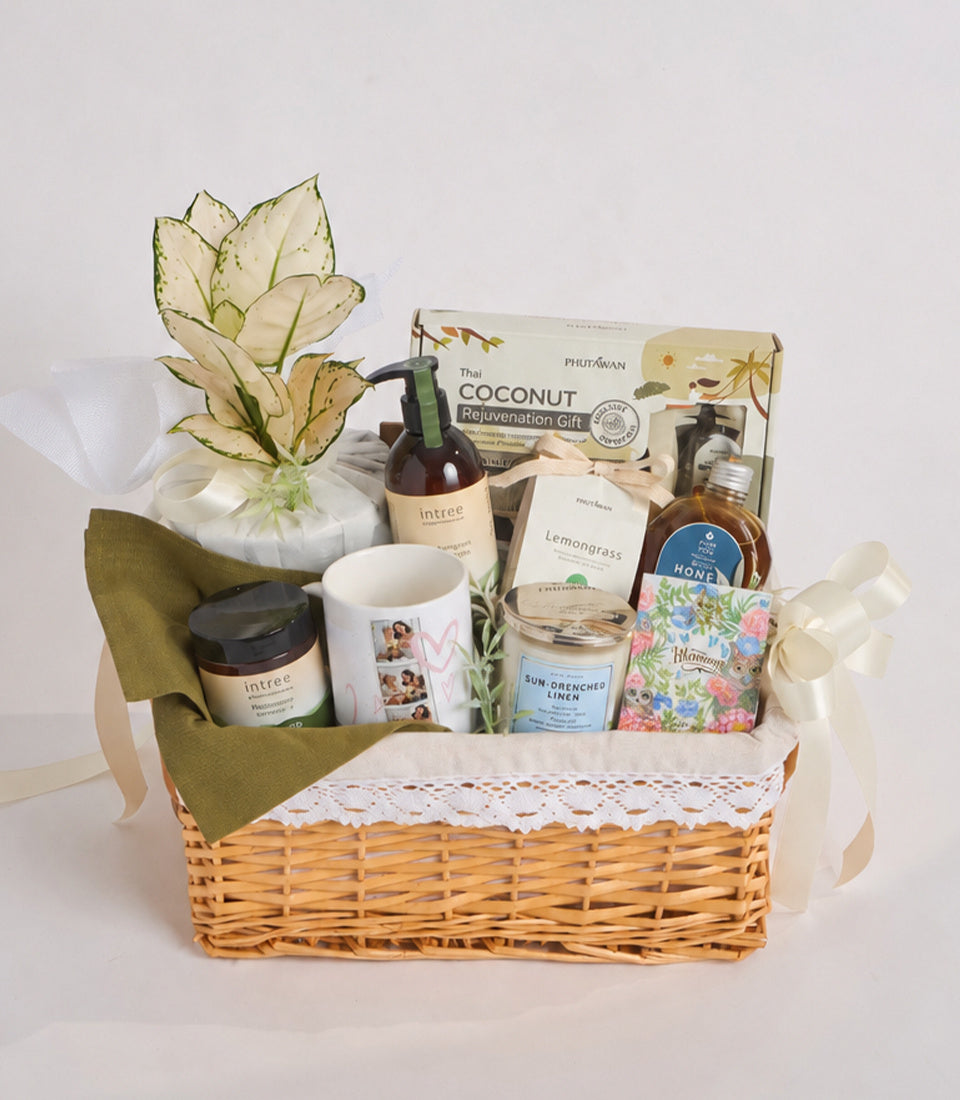 Wicker basket gift set with natural products, cream ceramic mug, glass jars, Intree skincare, honey, lemongrass candle, and decorative dried flower arrangement with green ribbon.