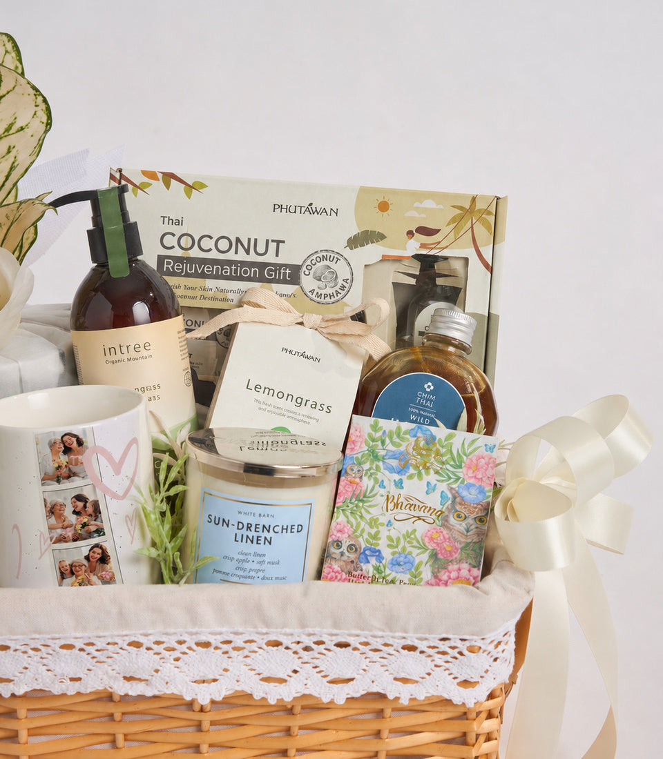 Wicker gift basket with white linen liner containing Thai spa products: coconut rejuvenation set, lemongrass soap, sun-drenched linen candle, organic mountain spray, and decorative photo card with cream ribbon bow.
