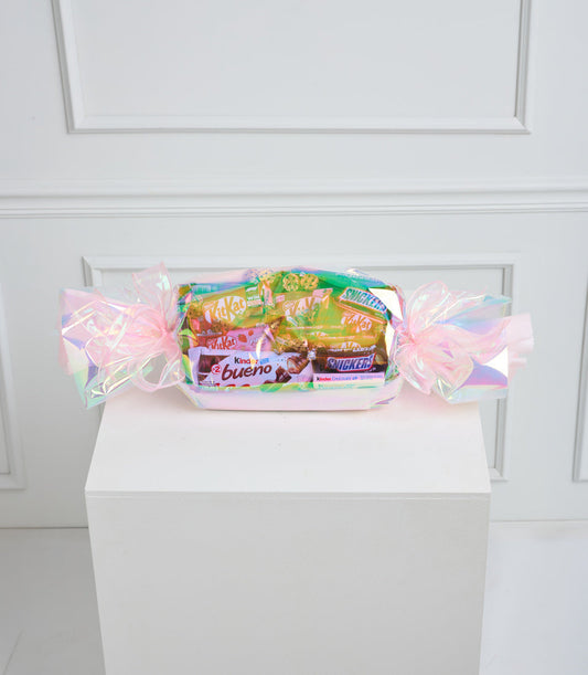 Assorted candy gift basket with pink iridescent cellophane wrapping and bows, filled with colorful chocolate and candy brands on white pedestal.