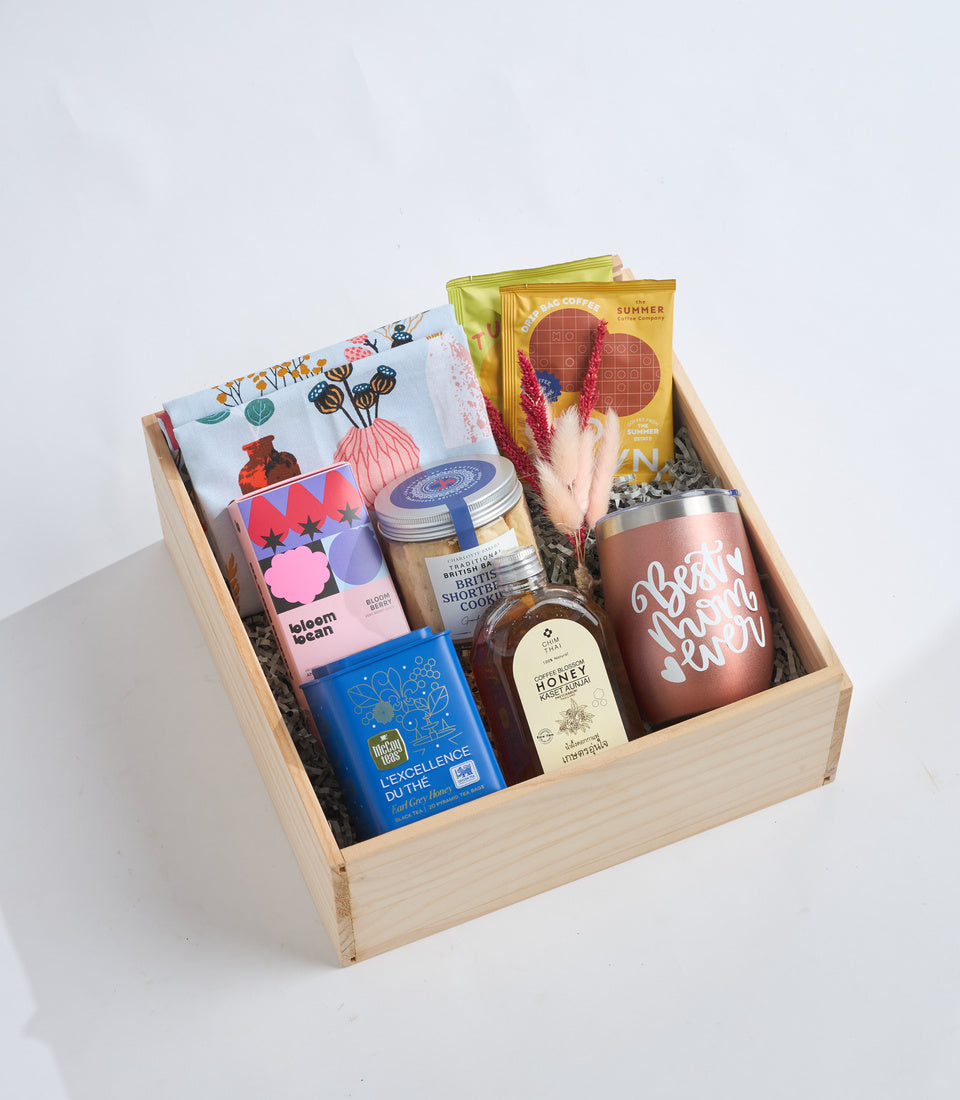 Wooden gift box with assorted items including floral tea towel, bloom band pouch, shortbread cookies, honey jar, coffee packet, dried flowers, and rose gold tumbler on white background.