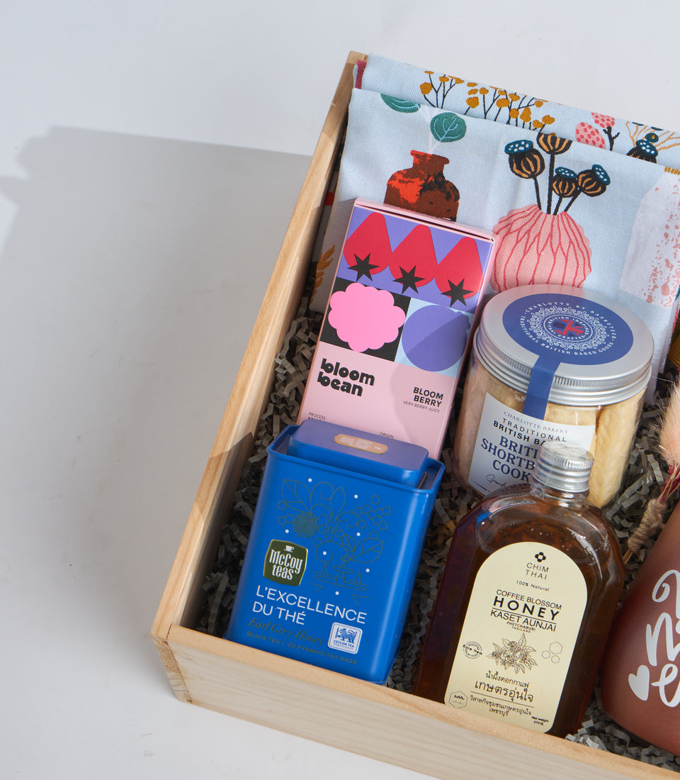 Wooden gift box with curated items: floral tea towel, bloom bean tea, McCoy Oolong tea, British shortbread cookies, and honey jars on shredded paper.