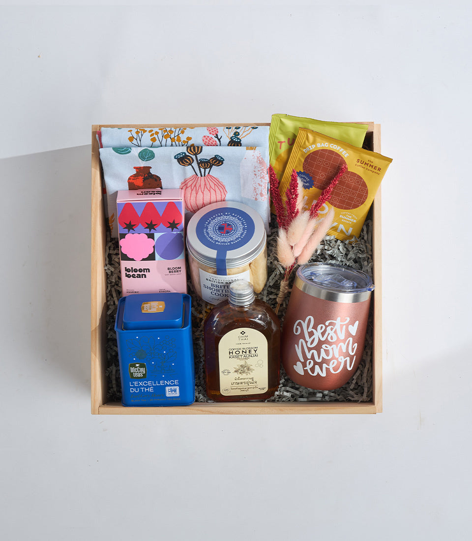 Curated gift box with natural wood frame containing skincare products, tea, honey, chocolate, a pink mug, dried flowers, and a floral patterned cloth on white background.