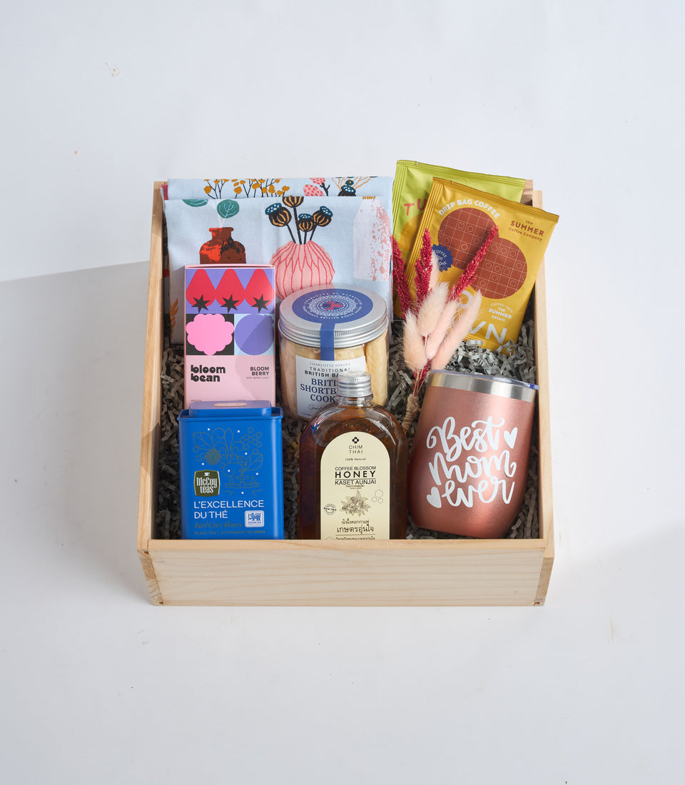 Wooden gift box containing colorful tea, honey, cookies, coffee bags, dried flowers, and a rose gold "Best Mom Ever" tumbler on white background.