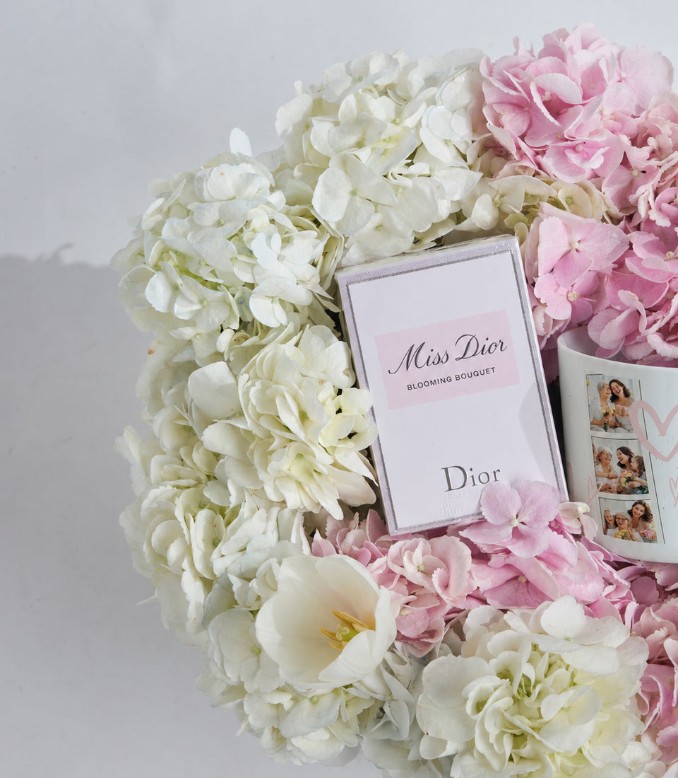 Miss Dior Blooming Bouquet perfume box surrounded by white and pink hydrangeas and peonies with photo prints on white background.