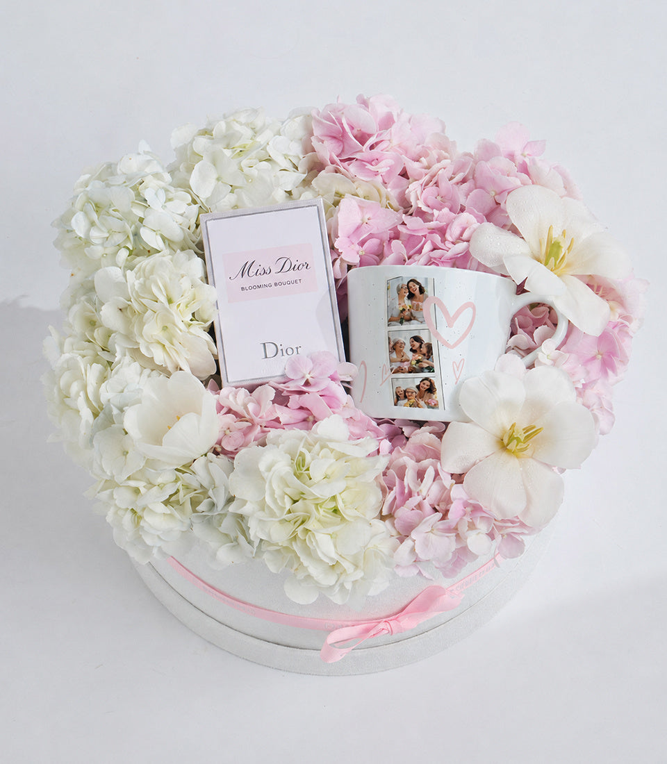 Luxury gift arrangement with white and pink peonies, Miss Dior perfume box, personalized mug with photos, and pink ribbon on white base.