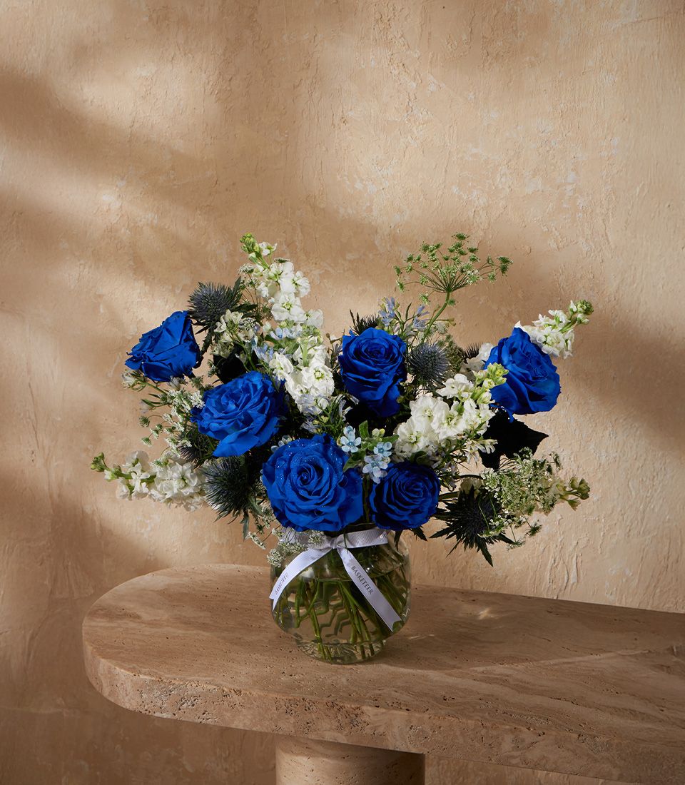 Deep blue roses and white stock flowers with thistle in clear glass vase with white ribbon bow