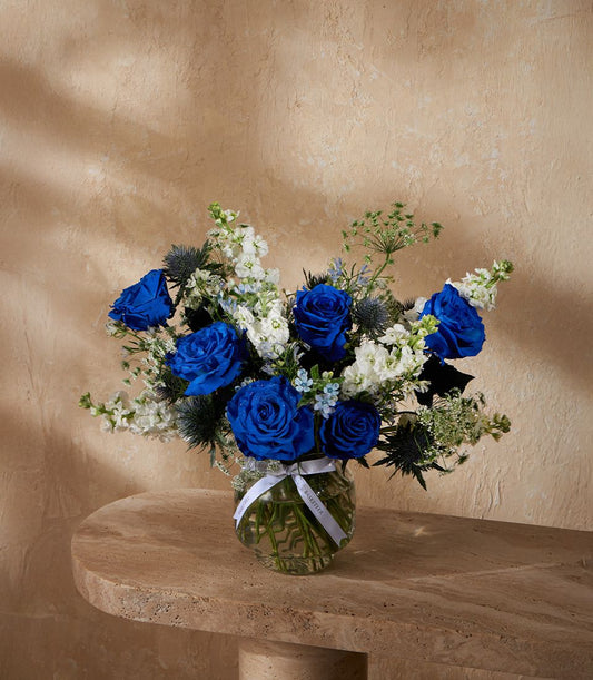 Deep blue roses and white stock flowers with thistle in clear glass vase with white ribbon bow