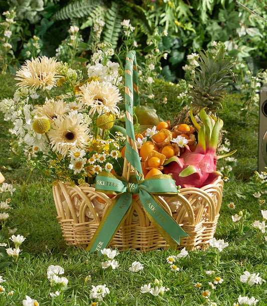 Wicker basket with tropical fruits, cream gerbera daisies, and white flowers tied with green gold-edged ribbon on garden lawn