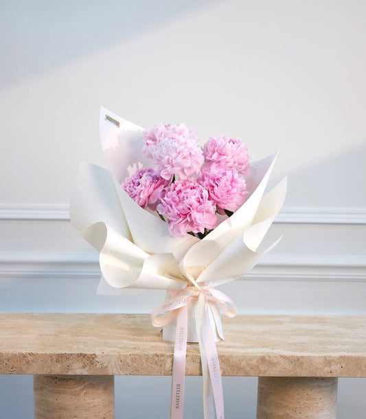 # Alt Text

Luxury bouquet of pink peonies wrapped in white paper with pale pink ribbon bow, displayed on natural stone shelf against white paneled wall.