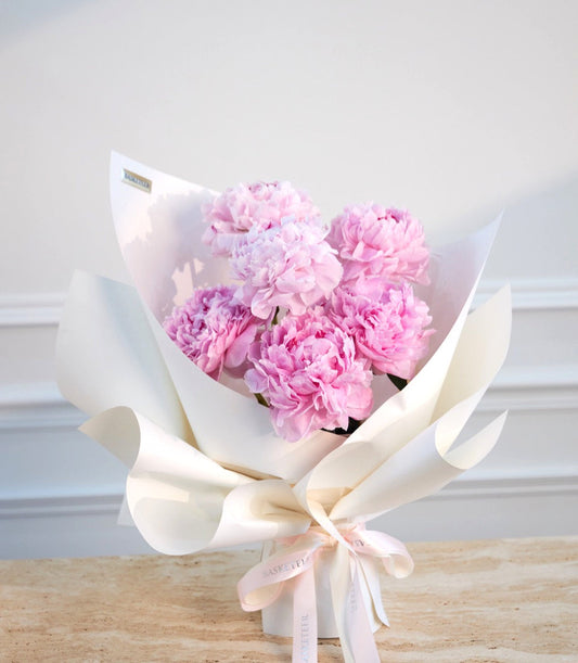 Pink peonies bouquet wrapped in white paper with cream ribbon bow, displayed on wooden surface against white background.