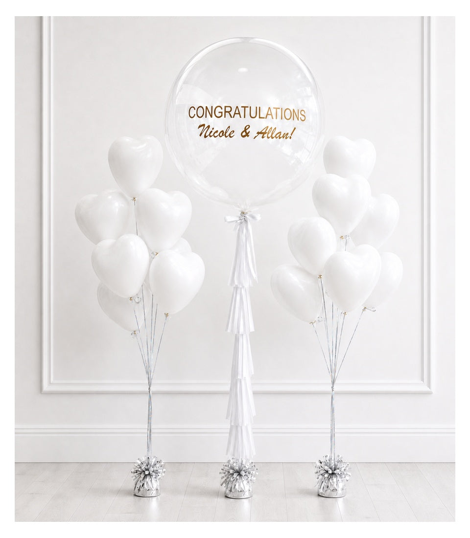 Elegant white balloon arrangement with oversized clear bubble balloon, personalized congratulations text, silver tassels, and weighted bases on light floor.