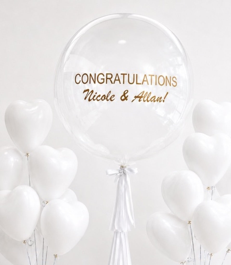 Clear bubble balloon with gold "Congratulations Nicole & Allan!" text, surrounded by white latex balloons with silver ribbons on white background.