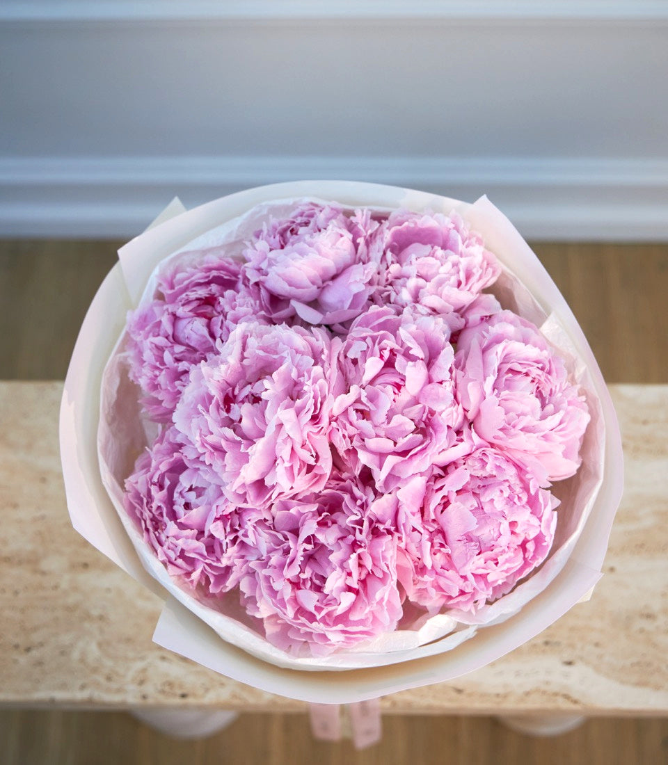 Pink peonies in white paper wrapping, arranged in a rounded bouquet with full, layered petals against a neutral background.