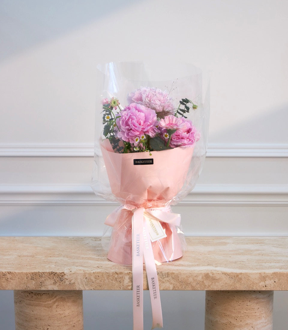Pink peony bouquet with clear cellophane wrapping, pink ribbons, and "Basketeer" florist label on cork-textured surface.