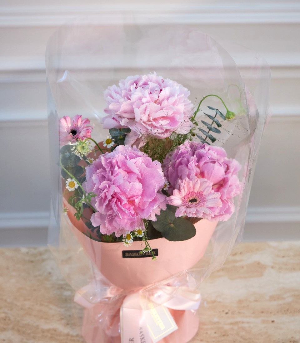 Pink peony and gerbera daisy bouquet in coral vase, wrapped in clear cellophane with pink ribbon bow.