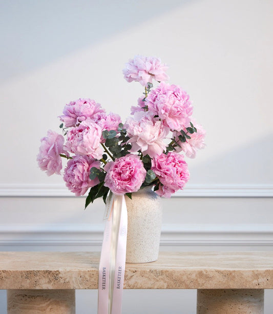 Pink peonies in cream concrete vase with pale pink ribbon bow on wooden shelf against white wall.