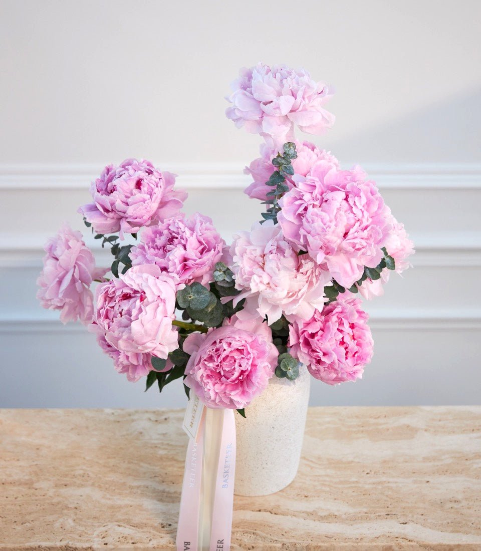 Pink peonies in cream ceramic vase with pink ribbon bow on wooden surface against white wall.