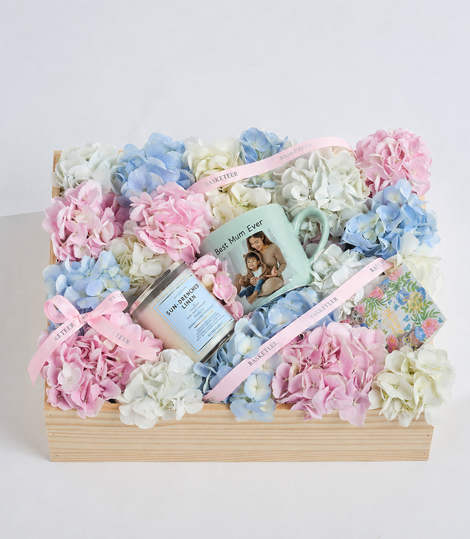 Wooden box arrangement with pink, blue, and white hydrangeas, skincare products, a photo mug, and pink ribbon bows on light background.