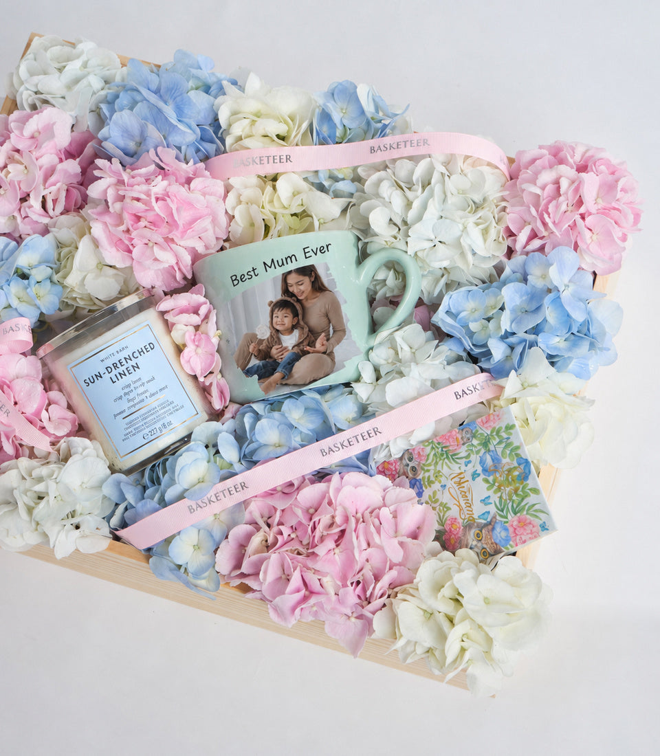 Heart-shaped floral gift basket with pink, blue, and white hydrangeas, personalized mug, candle, and ribbon on wooden base.